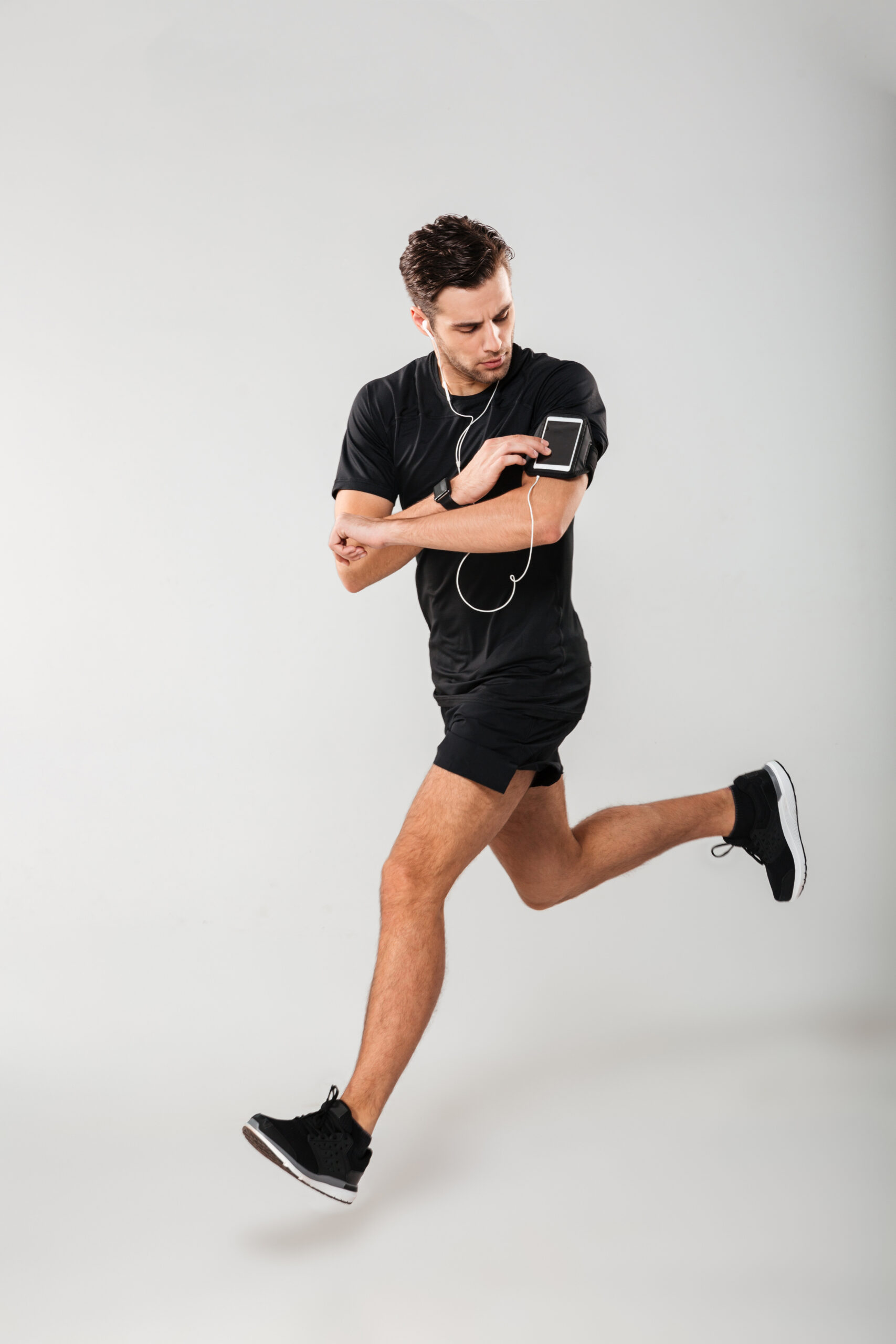Full length portrait of a handsome young man athlete in earphones listening to music while running and touching his phone in armband isolated over gray background